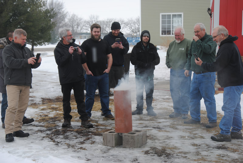 Men stand around a clay flue tile with smoke exiting the top and a vertical crack in the side of the tile.